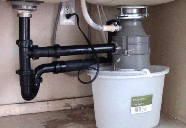 Under-sink view of a new garbage disposal installation with black pipes and a white bucket, showcasing a tidy plumbing setup.