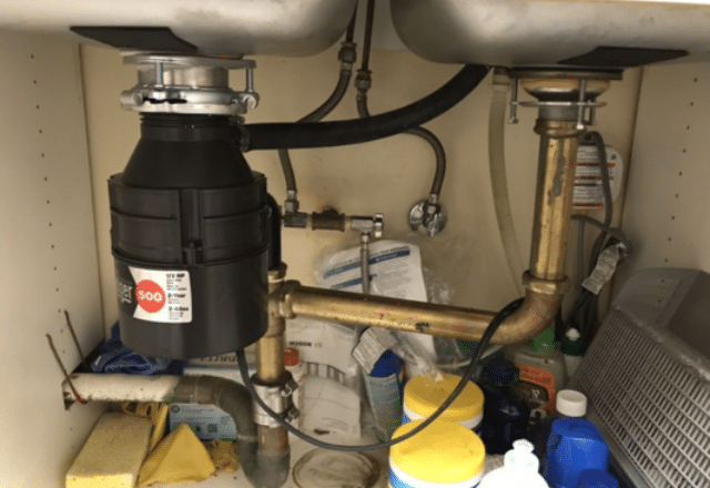 Under a kitchen sink with a newly installed garbage disposal, pipes, and a variety of household cleaning supplies.