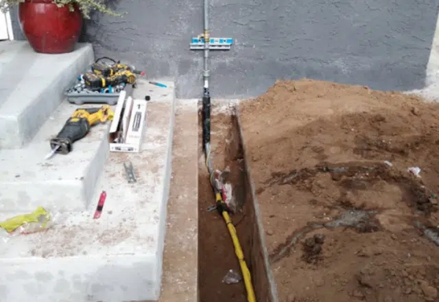 A trench revealing a new gas line installation along the side of a house, with a meter and tools visible.