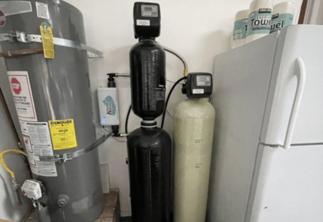 A water softener system installed next to a household water heater and refrigerator, showcasing components of a residential water maintenance setup.