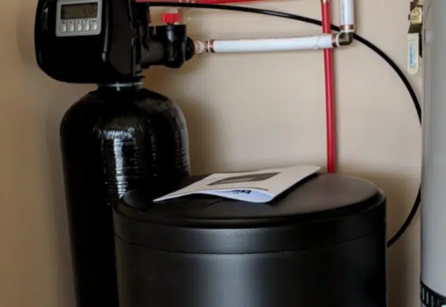 A close-up view of a water softener's control valve and connections, with a manual on top, as part of routine water softener maintenance.