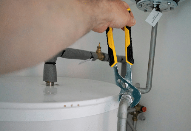 A hand using an adjustable wrench on a water heater's piping, representing the type of emergency plumber services needed for hot water issues