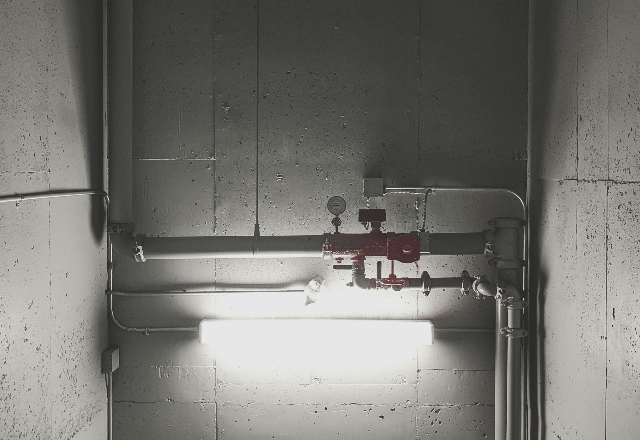 Industrial setting with PVC pipes and a lit fluorescent light fixture on a gray concrete wall, a scene a plumber in San Jose might work in
