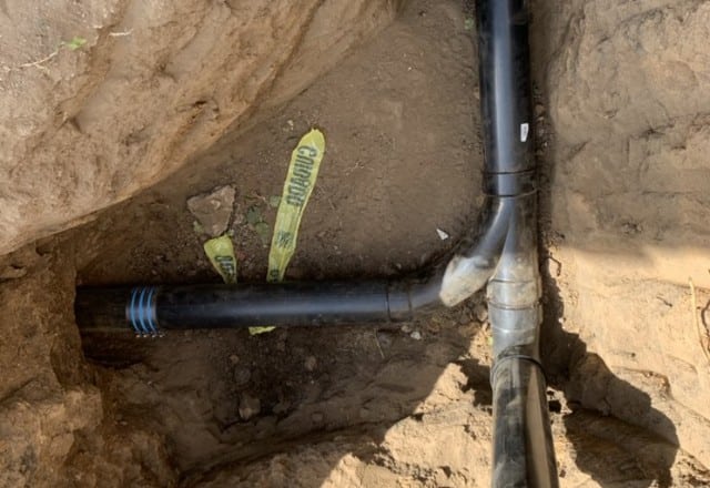 A black sewer pipe with connection to a solid black pipe, lying in a dirt trench, part of a larger sewer line replacement project
