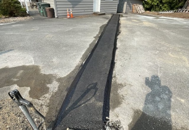 Freshly applied asphalt patch over trench for new sewer line installation, with shadow of a person and a camera tripod visible, highlighting recent sewer line replacement work