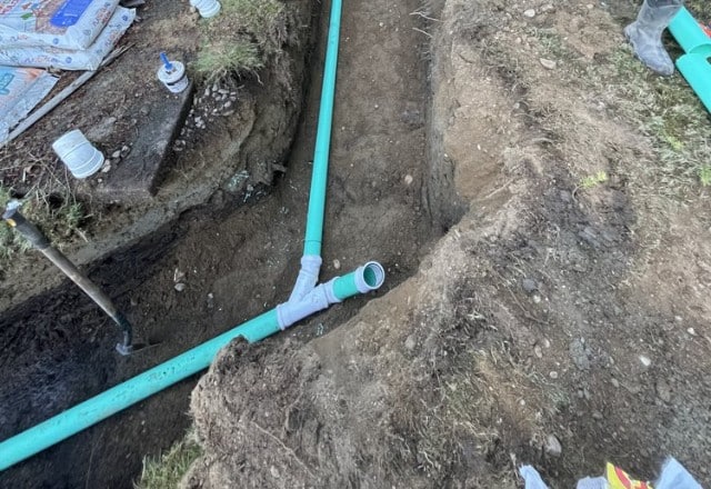 Trench revealing new green PVC sewer pipes with fittings and open ends, amidst brown earth, during a sewer line replacement operation.