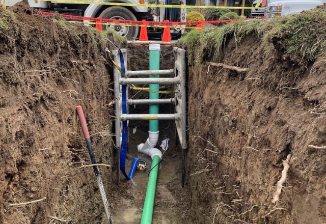 Deep trench with green sewer pipes and shoring equipment in place, displaying safety precautions taken during sewer line replacement.