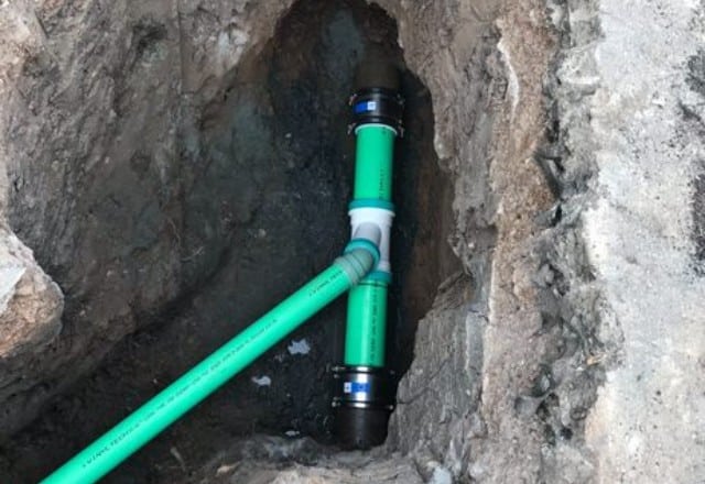 Green PVC sewer pipe with a black coupling, installed in a narrow trench with rocky soil, signifying part of a sewer line replacement process