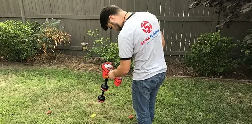 Plumber using a red electronic leak detection device in a grassy residential yard.